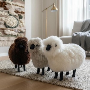 May include: Three sheep-shaped footstools with fluffy, textured wool-like upholstery in dark brown, light gray, and white. Each stool features four sturdy black legs and a pointed black snout, standing side-by-side on a textured area rug.