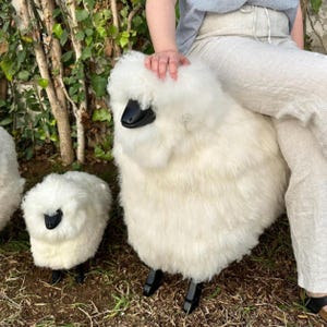 May include: White sheep-shaped ottoman with black legs and a black face. The ottoman is covered in fluffy white material. A smaller sheep ottoman is also visible. The ottomans are placed on the grass.
