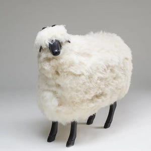 Könnte beinhalten: Ein schafförmiger Hocker mit einem dicken, flauschigen, cremeweißen Kunstfellbezug, der ein Schafsfell imitiert. Der Kopf ist schwarz und glatt, und die vier Beine sind ebenfalls schwarz, schlank und stabil, was dem Sitz eine feste Basis verleiht.
