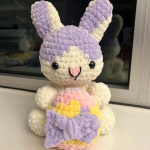 May include: A handmade crochet bunny plushie in white and lavender, holding a colorful Easter egg. The bunny has black safety eyes and a pink nose. The egg is yellow, pink, and lavender with a lavender bow. A cute Easter decoration.
