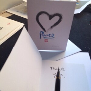 May include: A white greeting card featuring a black heart and the word "Peace" in blue. The inside of the card is blank, with the words "Thank you" being written with a pen.
