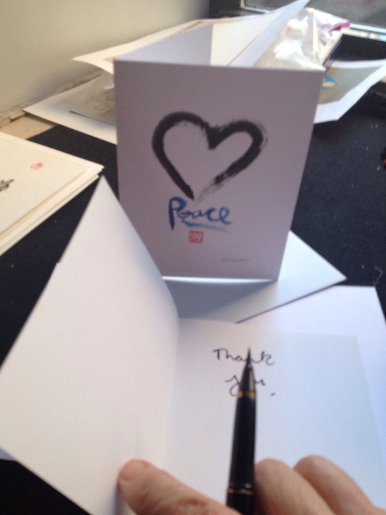 May include: A white greeting card featuring a black heart and the word "Peace" in blue. The inside of the card is blank, with the words "Thank you" being written with a pen.