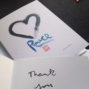 May include: A white greeting card featuring a black heart and the word "Peace" in blue calligraphy. The card is open, displaying the words "Thank you" in black script.