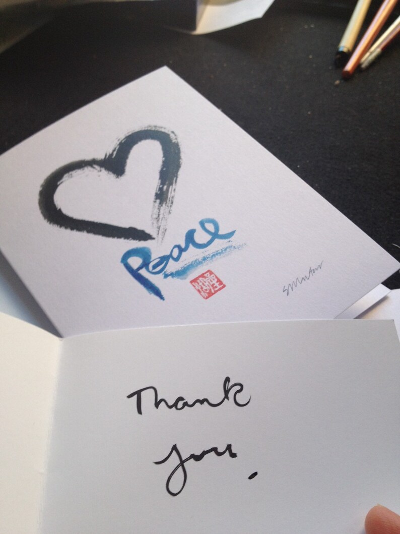 May include: A white greeting card featuring a black heart and the word "Peace" in blue calligraphy. The card is open, displaying the words "Thank you" in black script.