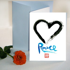 May include: A white greeting card featuring a black heart and the word "Peace" in blue script. A light blue envelope and an orange flower with green leaves are also present.