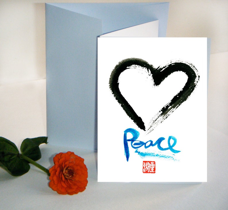 May include: A white greeting card featuring a black heart and the word "Peace" in blue script. A light blue envelope and an orange flower with green leaves are also present.