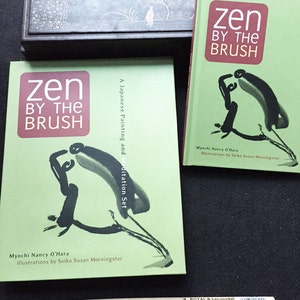 May include: Two books titled "Zen by the Brush" with green covers and abstract black ink illustrations. A wooden ink box and a calligraphy brush are also present. The books are by Myochi Nancy O'Hara, with illustrations by Seiko Susan Morningstar.