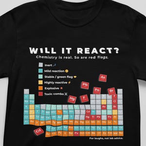 Op de afbeelding: Zwart T-shirt met de tekst "WILL IT REACT?" en een periodiek systeem ontwerp. Het shirt bevat een kleurgecodeerde sleutel voor chemische reacties, met zinnen als "Toxic combo" en "Explosive".