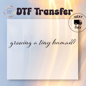 May include: White square with the text "growing a tiny human" in a cursive font, with a heart symbol. The words "DTF Transfer" are at the top. A "Next Day" delivery icon is on the right.