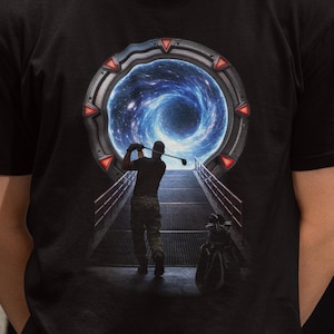 May include: Black t-shirt featuring a golfer swinging a club towards a swirling blue and white portal. The design includes a golf bag and a futuristic, sci-fi theme. The shirt is made of a dark fabric.