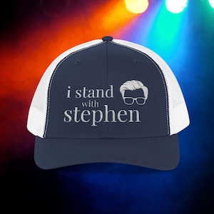 May include: Navy blue trucker hat with a white mesh back and a curved brim. The front of the hat features the embroidered text "i stand with stephen" and a graphic of a person's head with glasses.