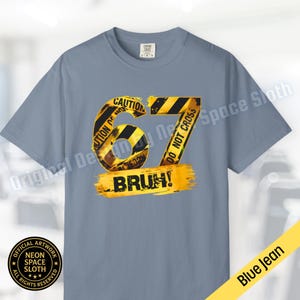 Puede incluir: Una camiseta azul vaquera con un diseño gráfico. El diseño presenta el número "67" en cinta de precaución amarilla y negra, con la palabra "BRUH!" en amarillo debajo. La camiseta tiene cuello redondo y mangas cortas.