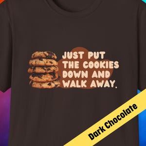 May include: A dark chocolate brown t-shirt with the text "Just put the cookies down and walk away." printed in white. A stack of chocolate chip cookies is pictured to the left of the text.