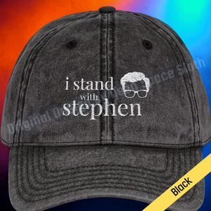 May include: Black, distressed-look baseball cap with the phrase "i stand with stephen" in white lettering. A white graphic of a person's head with glasses is to the right of the text. The cap has a curved brim and an adjustable strap.