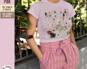 Può includere: Una maglietta rosa chiaro con una stampa floreale che presenta un bouquet di fiori in tonalità rosa, bianco e verde. La maglietta ha un collo rotondo e maniche corte. La modella indossa la maglietta con un paio di pantaloncini rosa a righe.