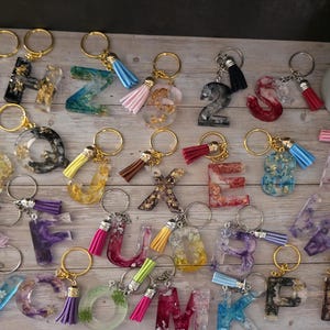 Könnte beinhalten: Eine Sammlung von Harz-Schlüsselanhängern in verschiedenen Farben, Formen und Designs. Jeder Schlüsselanhänger hat einen goldfarbenen Schlüsselring und eine Quaste. Die Schlüsselanhänger haben die Form von Buchstaben und Zahlen, mit dekorativen Elementen wie Goldflocken und Trockenblumen.