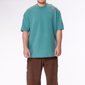 Op de afbeelding: Een turquoise t-shirt met korte mouwen en een klein geborduurd ontwerp op de linkerbovenborst, gecombineerd met een bruine werkbroek. De broek heeft meerdere zakken en een relaxte pasvorm. De outfit wordt gecompleteerd met zwarte sneakers.