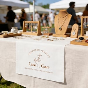 Puede incluir: Un puesto de joyería artesanal en un mercado al aire libre. Se exhiben collares, pendientes y pulseras en expositores de madera y un mantel blanco. El logotipo de Luna Grace es visible.