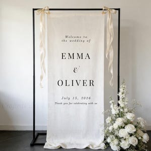 May include: A cream-coloured wedding welcome sign with the names EMMA & OLIVER and the date July 15, 2026. The sign is suspended from a black metal frame and decorated with cream ribbons. A floral arrangement of white roses and greenery is placed to the right of the sign.