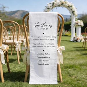 May include: White memorial chair sash with the text "In Loving Memory" and names of loved ones. The sash is draped over a wooden chair with a cream-colored bow. The background includes other chairs, an archway, and greenery.