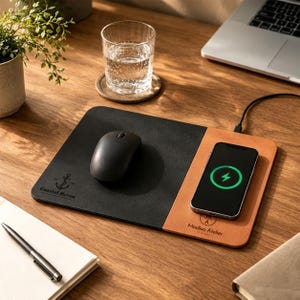 May include: A rectangular desk mat featuring a black surface on the left with an anchor logo and the text Coastal Haven, and a tan surface on the right with a wireless charging area, a black computer mouse, and a smartphone displaying a green icon.