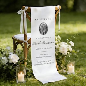 May include: A white satin chair sash with the word "RESERVED" at the top, a photo, and text that reads "In Loving Memory of Sarah Thompson, Mother of the Groom." The sash is draped over a wooden chair, with flowers and candles nearby.