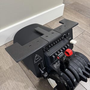 May include: Black aircraft control panel with multiple buttons, switches, and dials. The panel is mounted on a black base with a gear lever and a red button. The panel is on a wooden surface.
