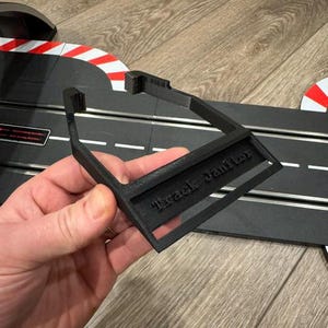 Könnte beinhalten: Ein schwarzer, 3D-gedruckter Streckenreiniger mit dem Text "Track Janitor" auf der Vorderseite. Der Reiniger wird vor einer Slotcar-Rennstrecke mit rot-weißen Streifen gehalten.