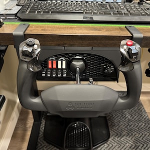 Puede incluir: Un yugo de simulador de vuelo gris con palancas de control y botones. El yugo está montado debajo de un escritorio de madera. La marca "Honeycomb Aeronautical" es visible en el yugo. Un teclado y un monitor están en el fondo.