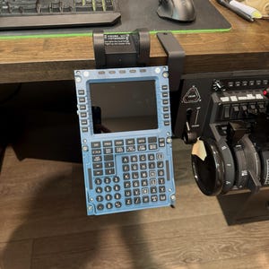 May include: A light blue flight simulator control panel with black buttons and a screen. The panel is mounted on a desk, near a keyboard, mouse, and throttle controls. The panel has an alphanumeric keypad and function buttons.