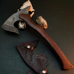 May include: A hand axe with a dark, weathered metal head and a curved, textured brown wooden handle secured with small brass pins. Beside the axe lies a dark brown leather sheath featuring intricate embossed patterns and two silver metal snaps.