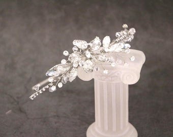 Può includere: Un accessorio per capelli color argento caratterizzato da una composizione floreale di cristalli trasparenti e piccole perline su una sottile struttura in filo metallico. Il design include cristalli a forma di foglia e delicati rametti.