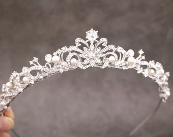 May include: A silver-toned metal tiara featuring an intricate filigree design adorned with clear sparkling rhinestones and small white pearls. The headpiece has a central floral motif and symmetrical scrolling patterns along the curved band.