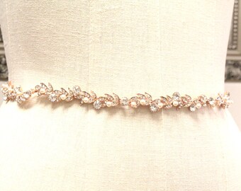 May include: A rose gold belt featuring a continuous vine-like design adorned with clear faceted crystals and small white pearls. The metallic structure is flexible and follows a repeating pattern of leaves and circular stones along its length.