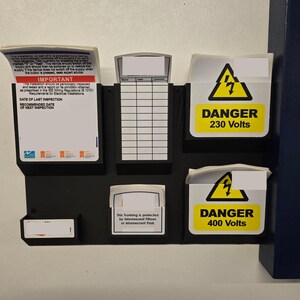 May include: A black electrical panel with several white and yellow warning labels. The labels read "DANGER 230 Volts" and "DANGER 400 Volts" with a lightning bolt symbol. There are also informational documents.