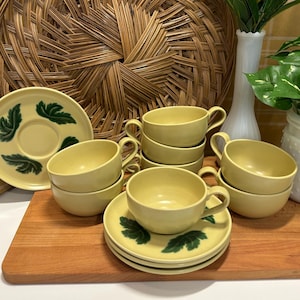 May include: A set of vintage yellow ceramic teacups and saucers. Each saucer features a green leaf design. The teacups are stacked, and the set is arranged on a wooden cutting board. A decorative wicker plate is in the background.