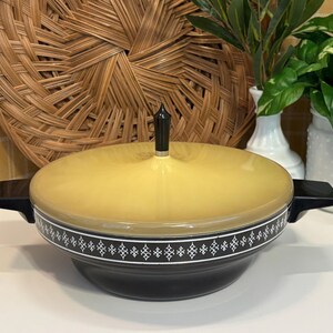 May include: A vintage yellow and black casserole dish with a lid. The dish has a black handle on each side and a decorative white and black band around the middle. The lid is topped with a black finial.