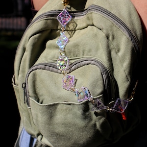 May include: An iridescent charm chain with diamond and cube shapes hangs from a gold clasp. The chain is attached to a green canvas backpack. The geometric shapes reflect multiple colours, creating a striking accessory.