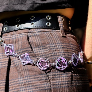 May include: A black leather belt with silver eyelets and a decorative chain belt with iridescent geometric shapes. The chain belt is draped over brown plaid trousers.