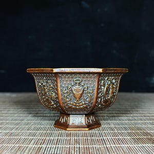 Może przedstawiać: Brązowa, ozdobna miska z sześciokątną podstawą i obramowaniem. Misa posiada szczegółowe rzeźbienia reliefowe z motywami kwiatowymi i geometrycznymi oraz centralny emblemat. Misa wykonana jest z ciemnobrązowego materiału i stoi na bambusowej macie.