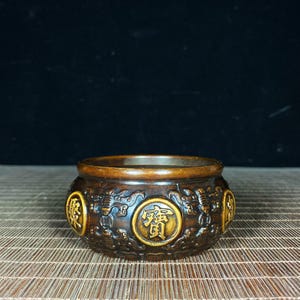 Może przedstawiać: Brązowy, okrągły palnik do kadzidła z misterzowskimi wzorami smoków i chińskich znaków. Naczynie ma ciemnobrązowy zewnętrzny i jaśniejszy złoty środek. Znaki znajdują się w okrągłym medalionie.