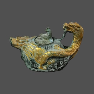 Könnte beinhalten: Eine verzierte Teekanne mit Drachendesign. Die Teekanne ist hauptsächlich aus Bronze mit goldenen Akzenten auf dem Kopf, dem Körper und dem Griff des Drachen. Der Deckel zeigt eine kleine vogelartige Skulptur. Der Boden der Teekanne hat chinesische Schriftzeichen.