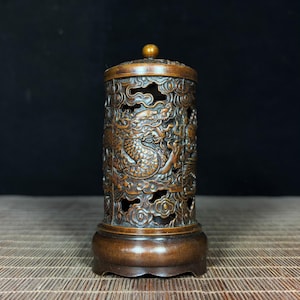 Może przedstawiać: Brązowy, cylindryczny palnik do kadzidła z misternymi rzeźbami smoków. Palnik ma zdejmowaną pokrywę z małym pokrętłem i stoi na okrągłej podstawie. Boki zdobią ażurowe wzory, pozwalające na ulatnianie się dymu z kadzidła. Całość jest szczegółowa.