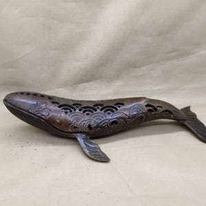 May include: A bronze whale-shaped decorative object. The whale features intricate wave-like patterns and cut-out designs along its back. The tail and fins are detailed with carved lines. The object has a dark, aged patina, suggesting it is vintage or antique.