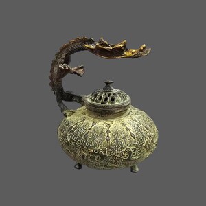 Könnte beinhalten: Ein antiker Weihrauchbrenner aus Bronze mit einem Drachengriff. Der runde Körper ist mit geätzten Schriftzeichen bedeckt und ruht auf drei kleinen Füßen. Der Deckel hat eine Reihe kreisförmiger Öffnungen. Der Drachenkopf ist goldfarben.