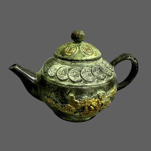 Könnte beinhalten: Eine antike Bronzeteekanne mit dunkelgrüner Patina. Die Teekanne weist Münz- und Drachenmotive auf, mit einem kleinen Knopf auf dem Deckel. Der Griff und der Ausguss sind dunkel und kontrastieren mit dem helleren grünen Körper und goldenen Akzenten.