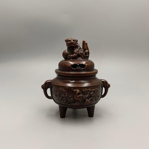 May include: Bronze incense burner with a detailed design. The burner features a lid with a small lion figure on top. The body of the burner has handles and is decorated with relief carvings. The burner stands on three legs.