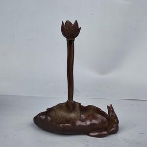 May include: A bronze-coloured lotus flower candle holder. The candle holder features a lotus flower at the top of a slender stem, rising from a detailed lotus leaf base. The sculpture is made of metal and has a detailed design.