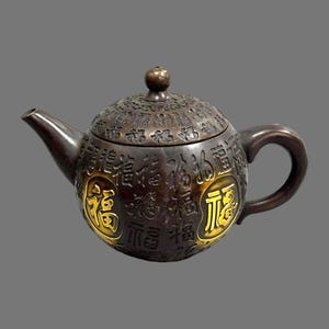 Könnte beinhalten: Eine bronzefarbene Teekanne mit rundem Körper, Ausguss und Henkel. Der Deckel und der Körper sind mit chinesischen Schriftzeichen graviert, wobei zwei Zeichen goldfarben hervorgehoben sind. Die Teekanne hat einen kleinen, runden Knopf auf dem Deckel.