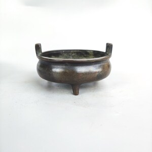 May include: A bronze-coloured, three-legged incense burner with two small handles. The bowl-shaped vessel has a dark patina, suggesting age. The interior shows a darker hue, possibly from use. The overall design is simple and functional.